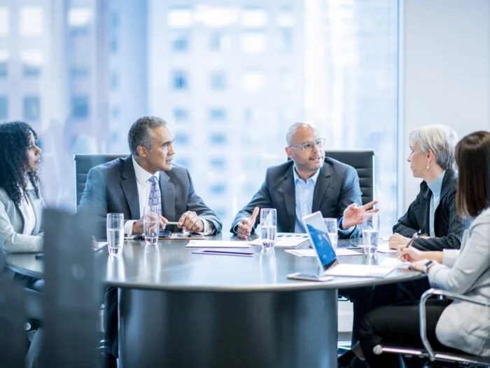 A diverse group of professionals engages in a meeting around a modern conference table, with laptops and documents present.