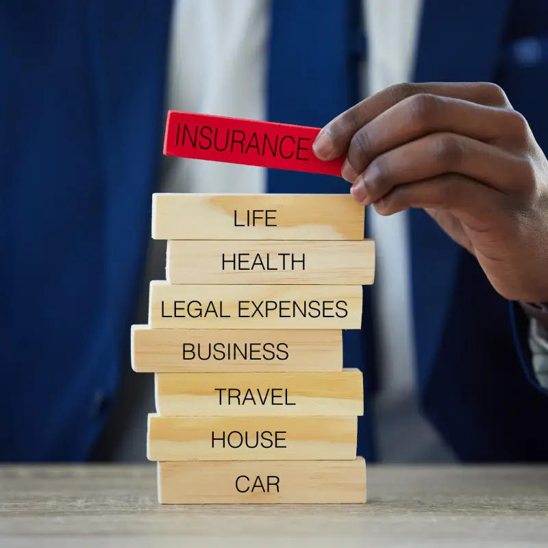 A person in a suit stacks wooden blocks labeled with various types of insurance, placing a red "INSURANCE" block on top.