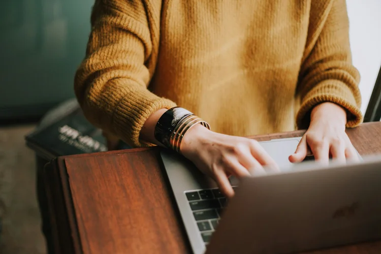 A close-up of a person's hands typing on a laptop, wearing a cozy mustard sweater and stylish bracelets, with a wooden desk in the background.